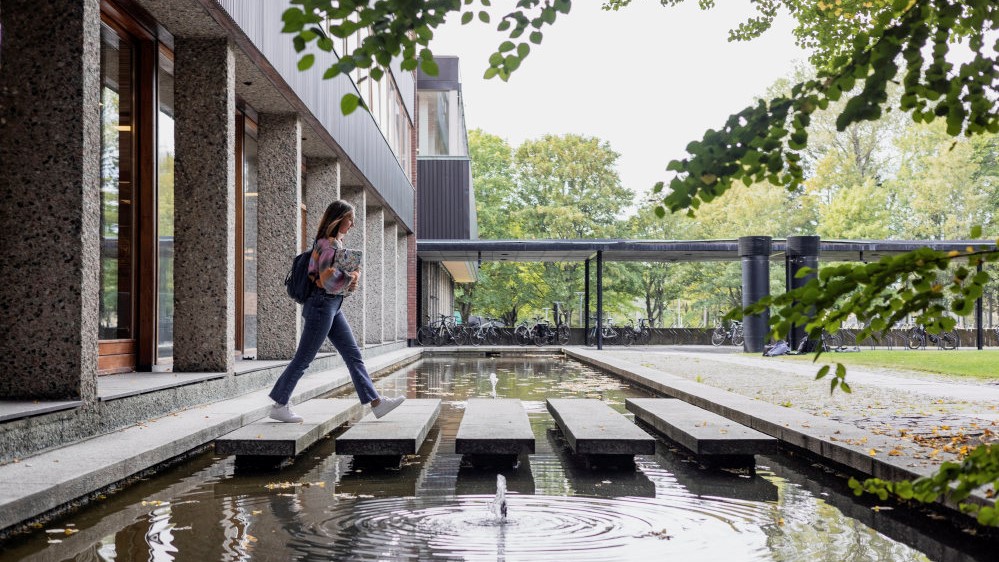 En student med bøker i hånda som går på fem trinn over et smalt vannspeil.