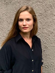 Bildet kan inneholde: kvinne, profil, smilende, langt brunt hår, mørk blå skjorte, grå mur i bakgrunn