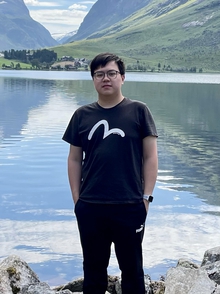 Ung mann star foran en innsjø, dal og fjell i bakgrunnen, svart kort hår, briller, sort t-skjorte, sorte bukser