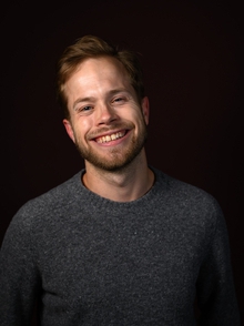 Portrettfoto, mann, stort smil, kort blondt hår, grå genser, sort bakgrunn