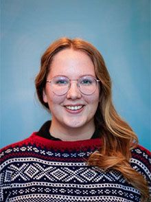 portrait photo of smiling woman, blue background, reddish long hair, dark blue sweater with white and red pattern