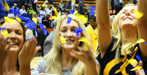 Tre blonde studiner smiler og kaster gul og blå konfetti opp i luften. Foto.